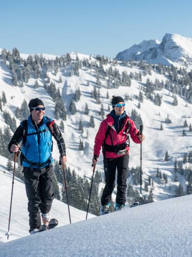 Zwei Tourenski-Wanderer vor einem schneebedeckten Hügel sind zu sehen.