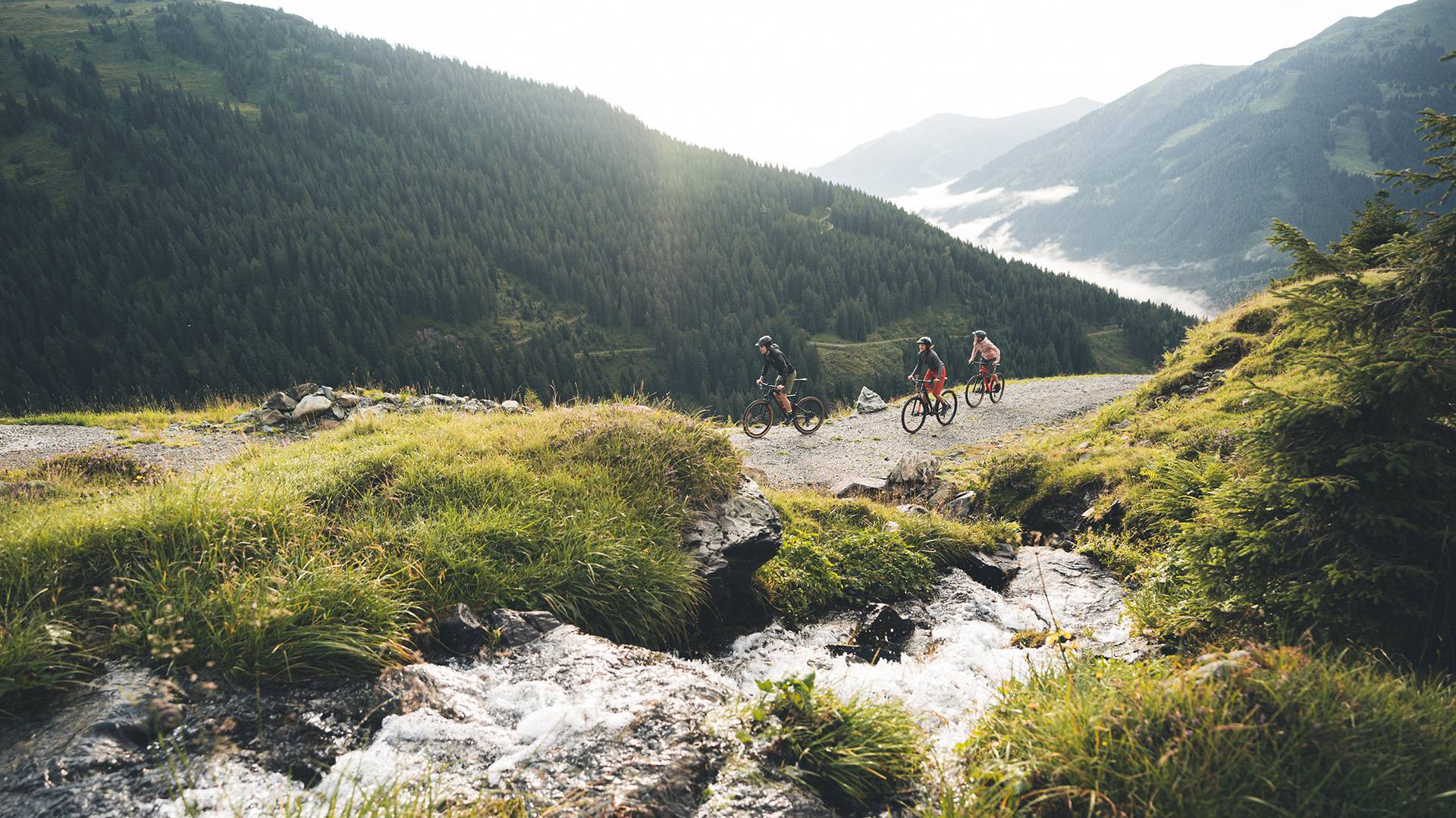 Drei Biker fahren neben einem Bach einen Schotterweg den Berg hinauf.