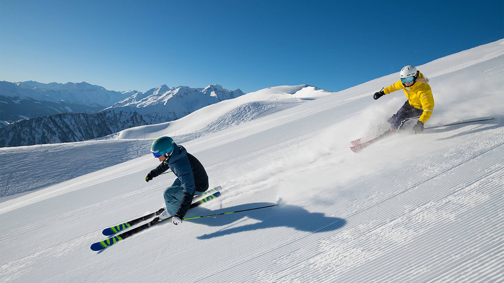 Zwei Personen carven auf einer perfekt präparierten Piste in Richtung Tal und haben dabei sichtlich Spaß.