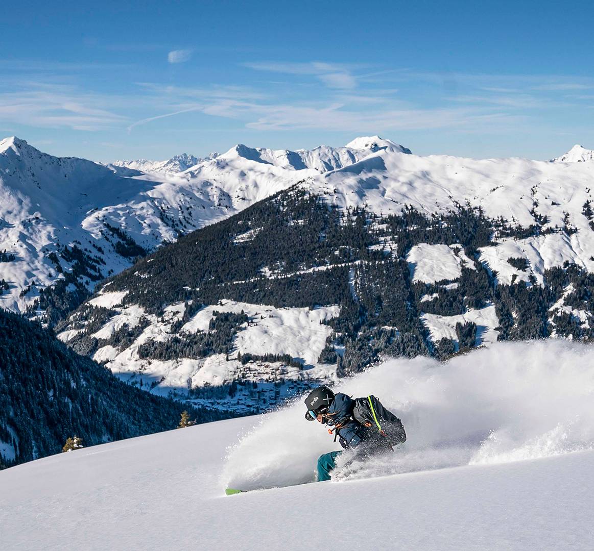 Ein Skifahrer fährt mit hohem Tempo die Piste hinunter und wirbelt dabei Schnee hinter sich auf.