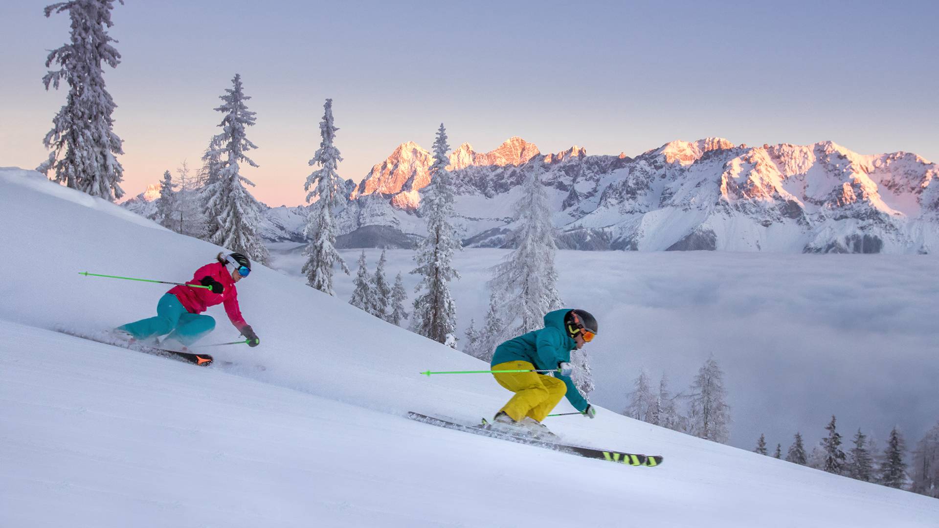 Two people ski down the slope one behind the other at sunset.