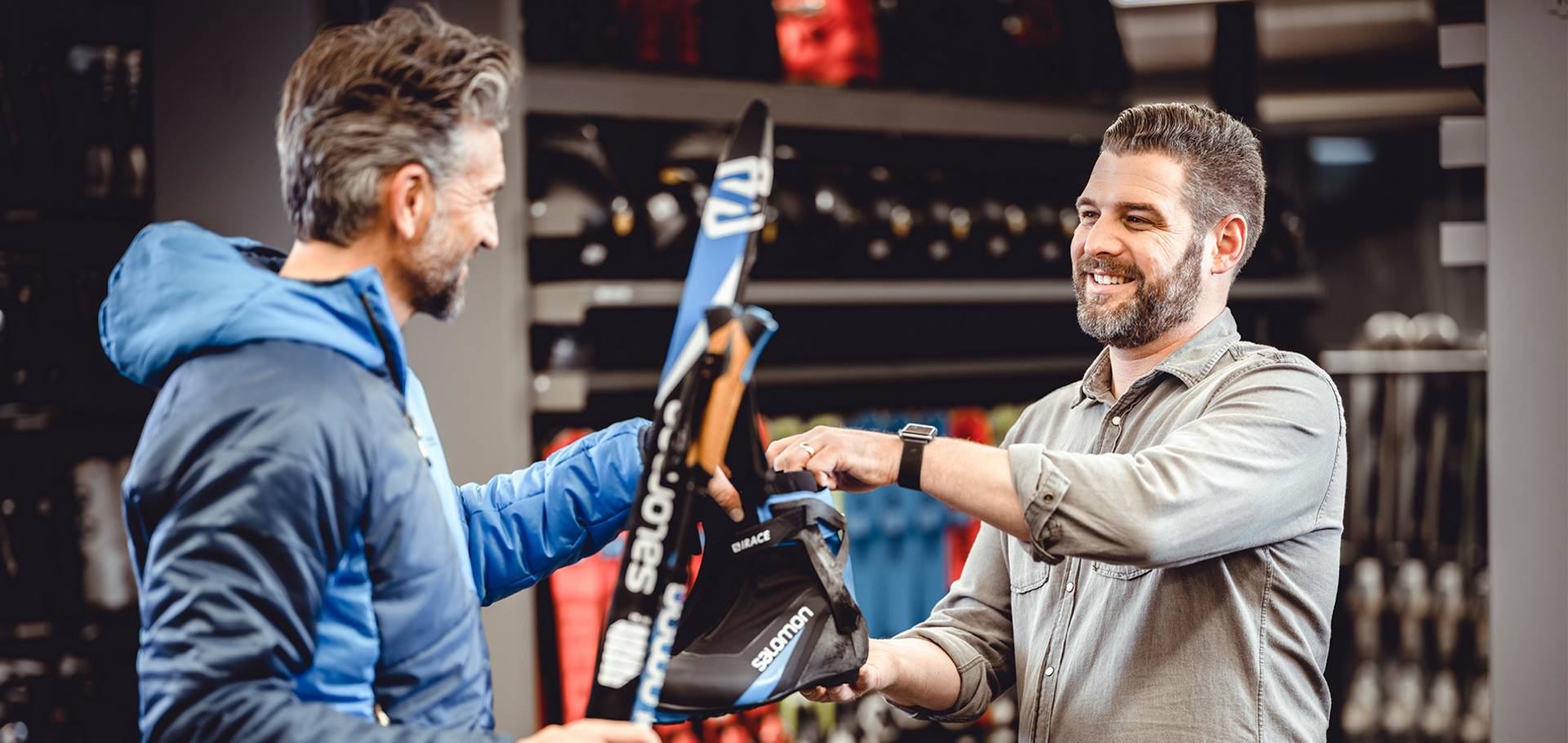 Een medewerker reikt een klant, die al ski's vasthoudt, skischoenen aan.