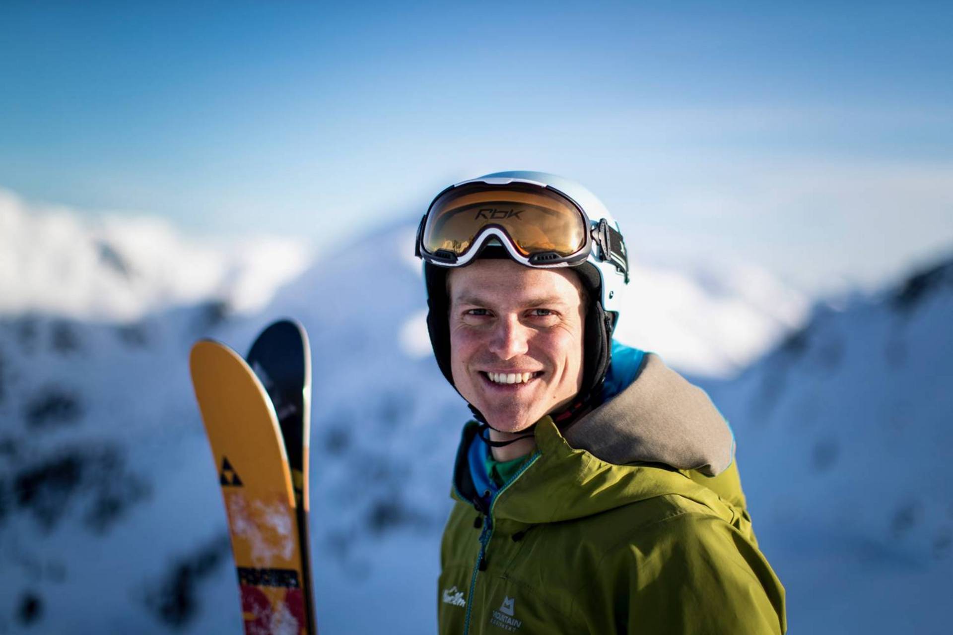 A skier beams at the camera in a close-up. He is holding his skis in his hand and in the background is the beautiful mountain panorama of the Planneralm.