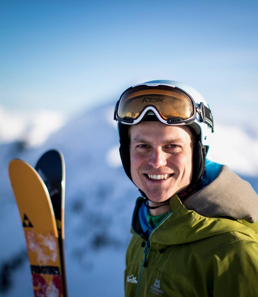 Ein Skifahrer strahlt in einem Close-up in die Kamera. In der Hand hält er seine Ski und im Hintergrund das wunderschöne Bergpanorama der Planneralm.