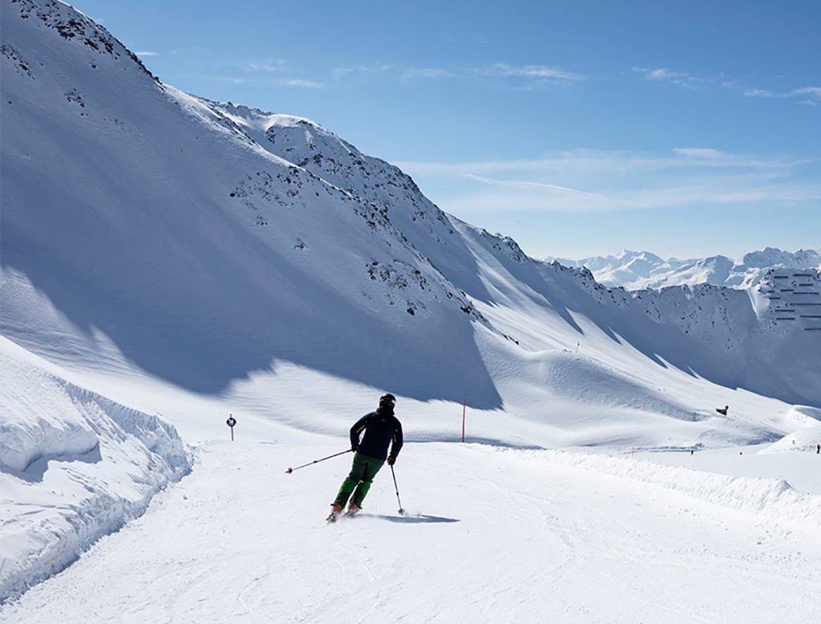 Ein einzelner Skifahrer zieht seine Schwünge auf der Piste, im Hintergrund ist ein Bergpanorama zu sehen.