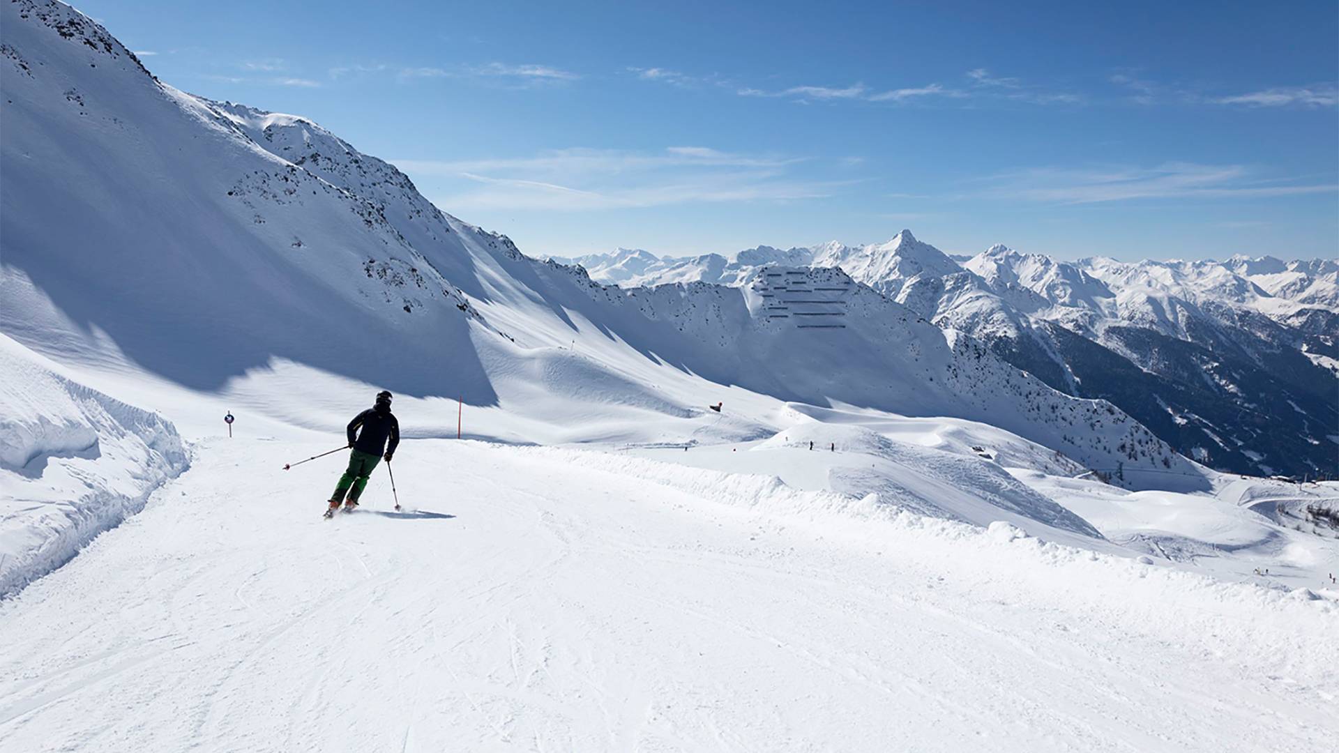 A single skier carves turns on the slope, with a mountain panorama in the background.