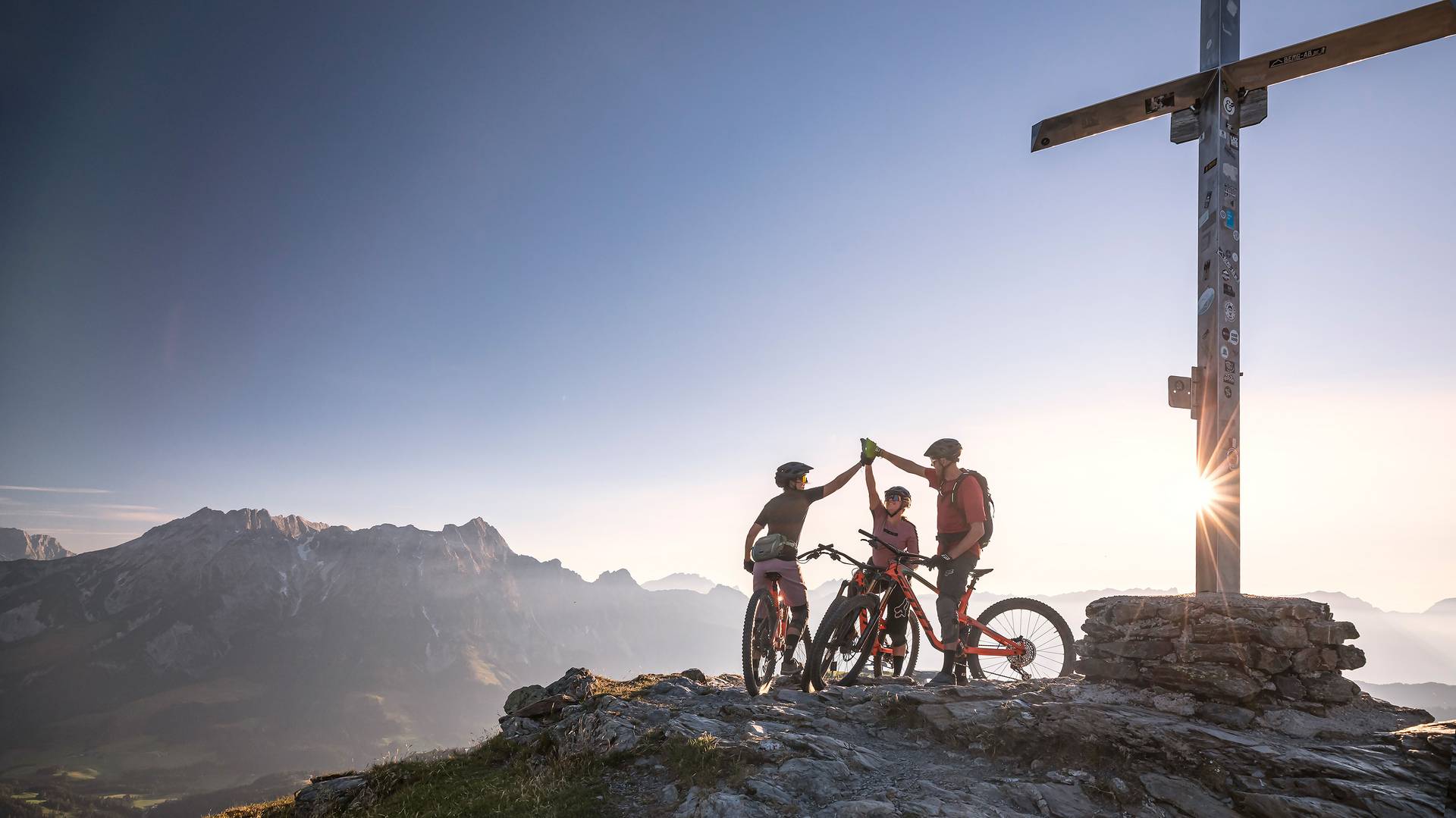 Drei Mountainbiker feiern mit einem ausgiebigen "high five" die geschaffte auffahrt zur Bergspitze während im Hintergrund der Sonnenuntergang das Bergkreuz erstrahlen lässt. 