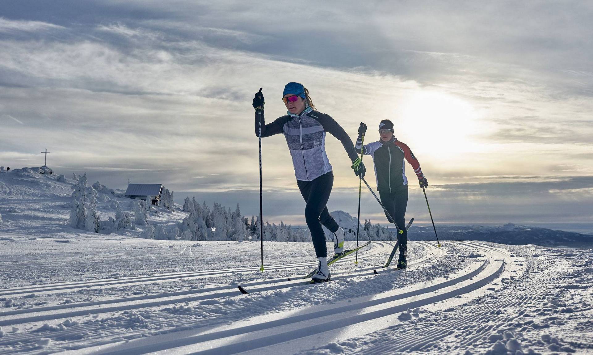 Two cross-country skiers can be seen skiing one behind the other on a trail in a dreamlike evening atmosphere.