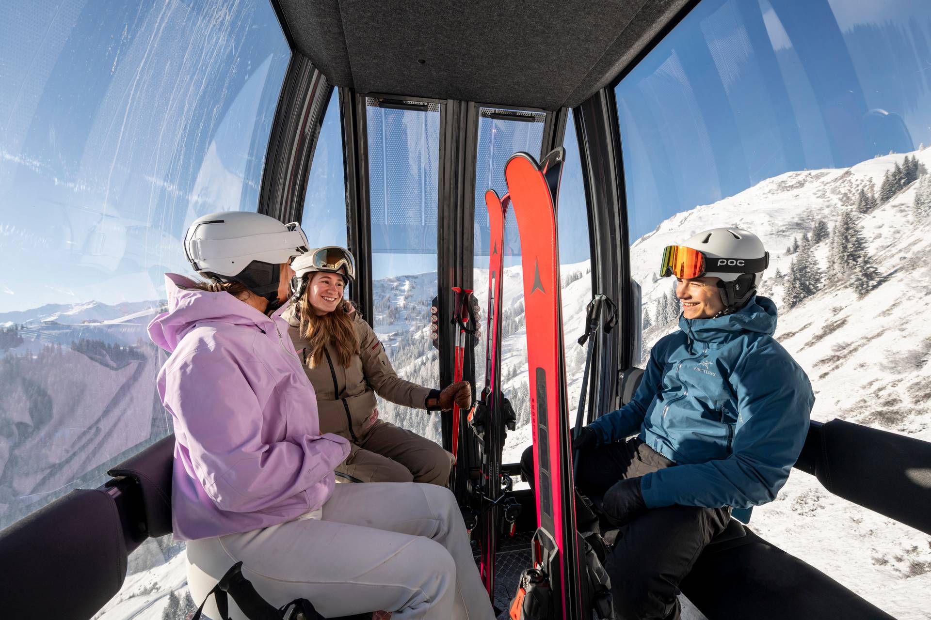 Drei Skifahrer sitzen in der Gondel mit Skiern, alle tragen einen Helm