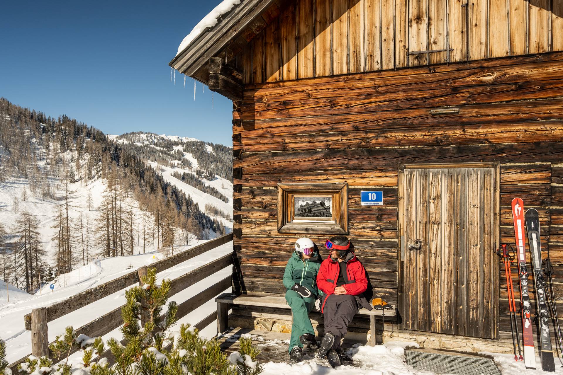 Pärchen sitzt vor einer Holzhütte im verschneiten Skigebiet Zauchensee, es schein die Sonne