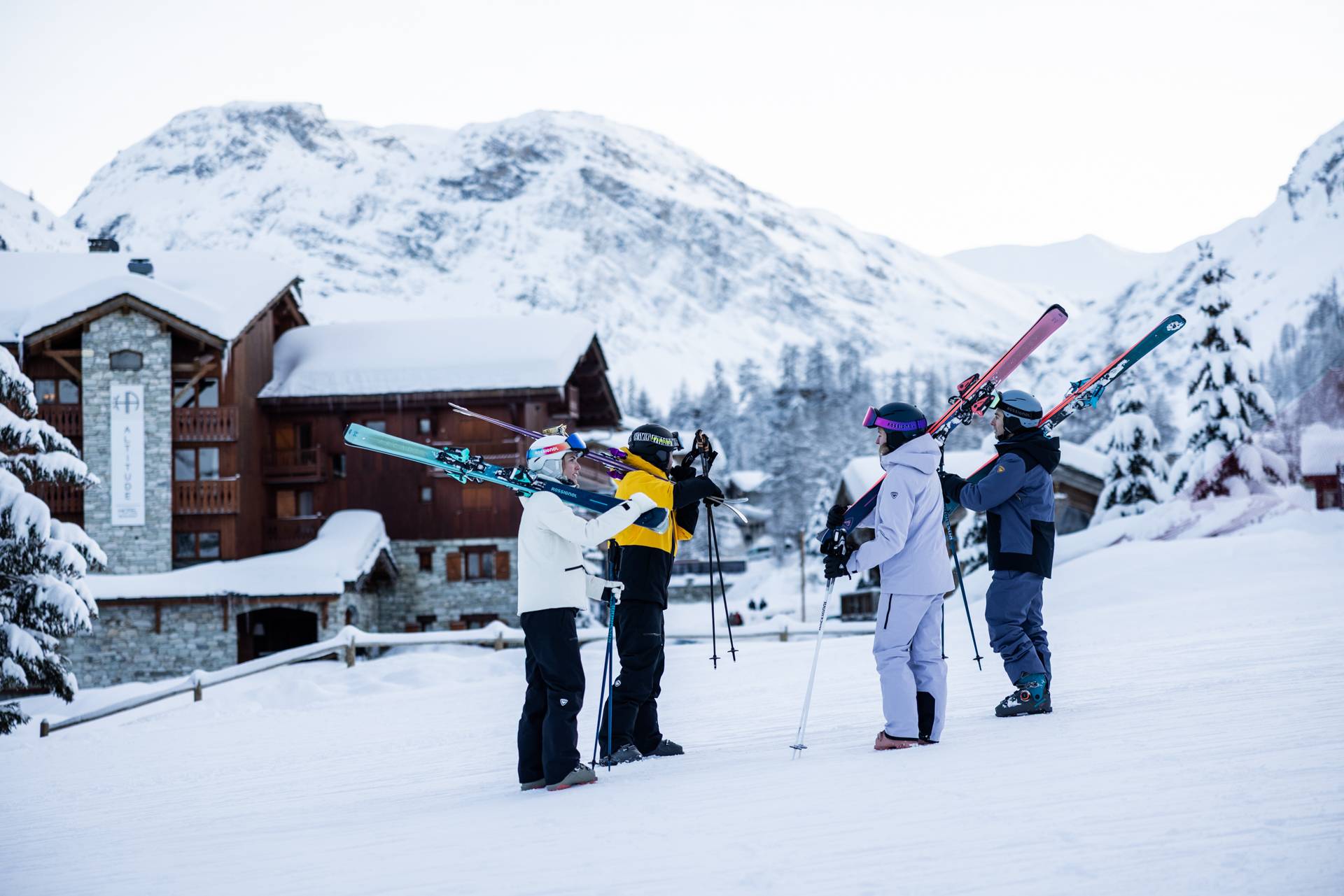 Een groep skiërs staat op de piste, ze dragen hun ski's op hun schouders. Op de achtergrond is een hotel te zien.