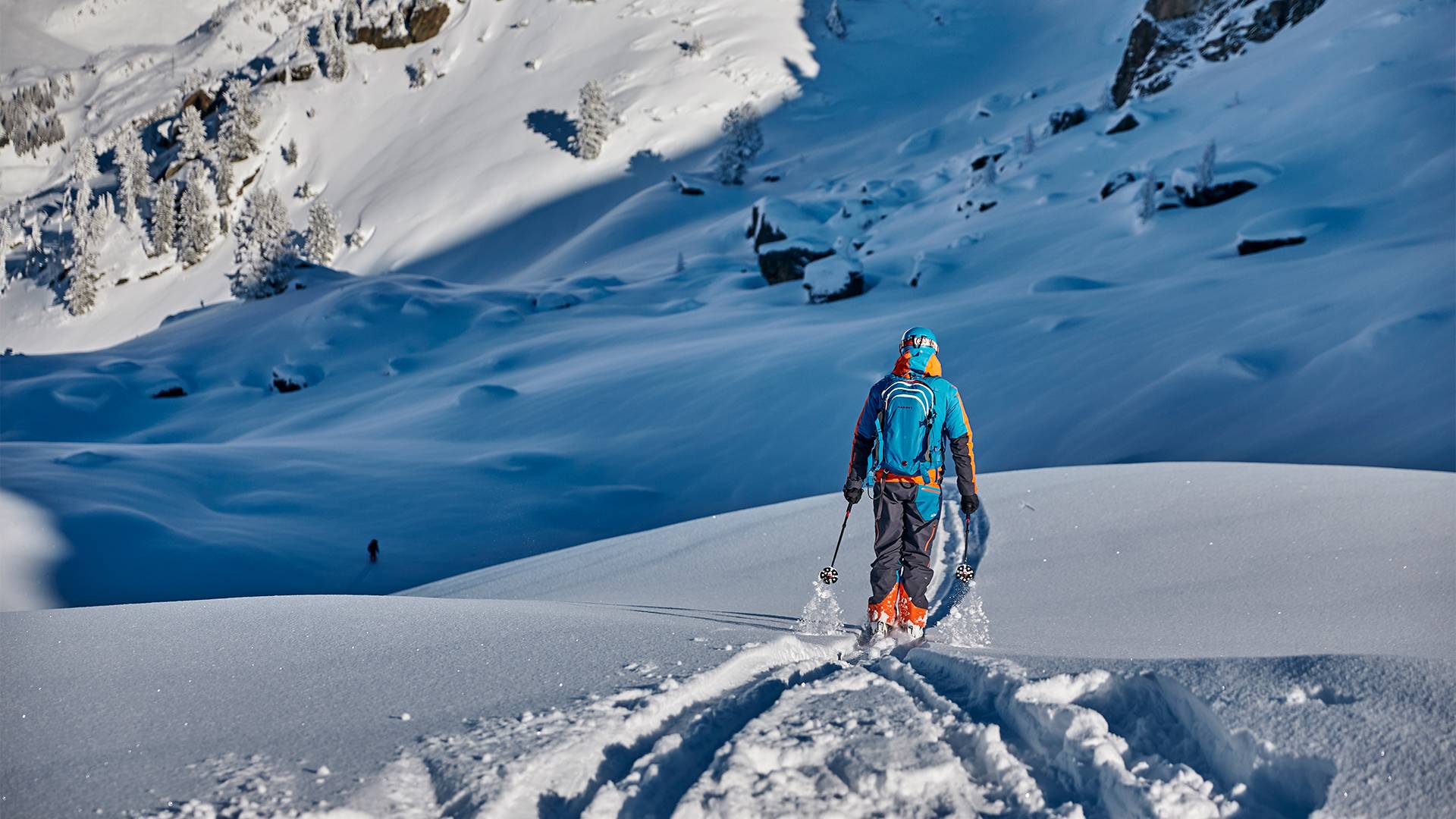 Ein Mann mit optimaler Winterausrüstung steht vor einem Neuschnee bedeckten Hang und freut sich darauf die Piste hinunter zu fegen.