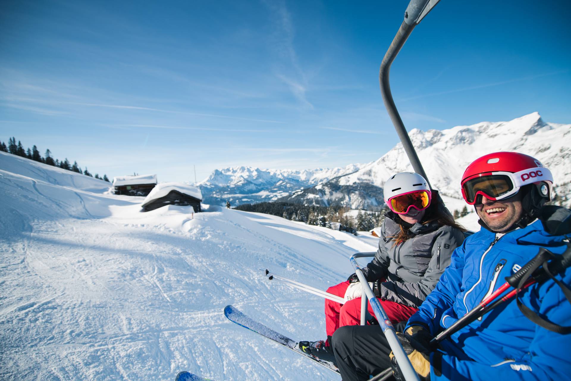 Two skiers are sitting on a chairlift, with a snow-covered hut visible in the background.