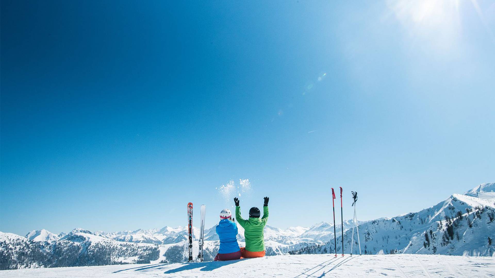 Two skiers sit off the piste and enjoy the sunny day and the view of the snow-covered mountain panorama