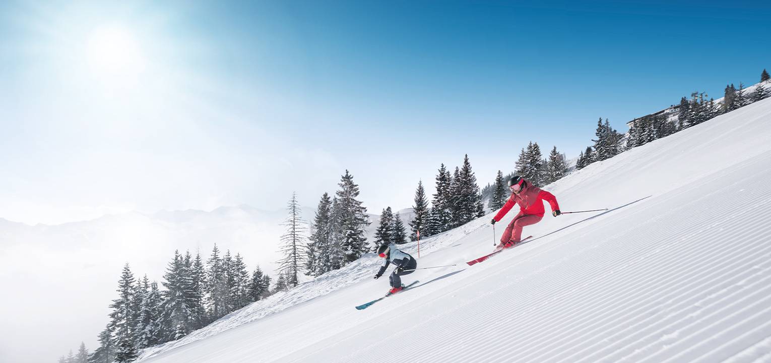 Zwei Skifahrer fahren auf der Piste, die Sonne scheint, im Hintergrund sieht man Nadelbäume