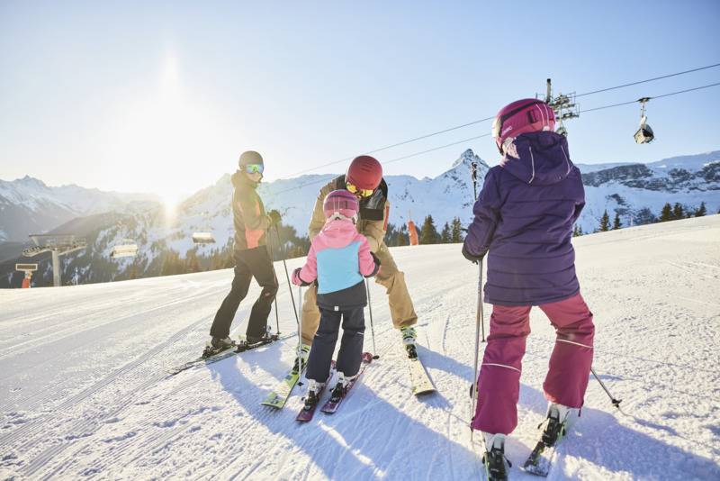 Familie steht auf der Skipiste, im Hintergrund scheint die Sonne und man sieht den Sessellift