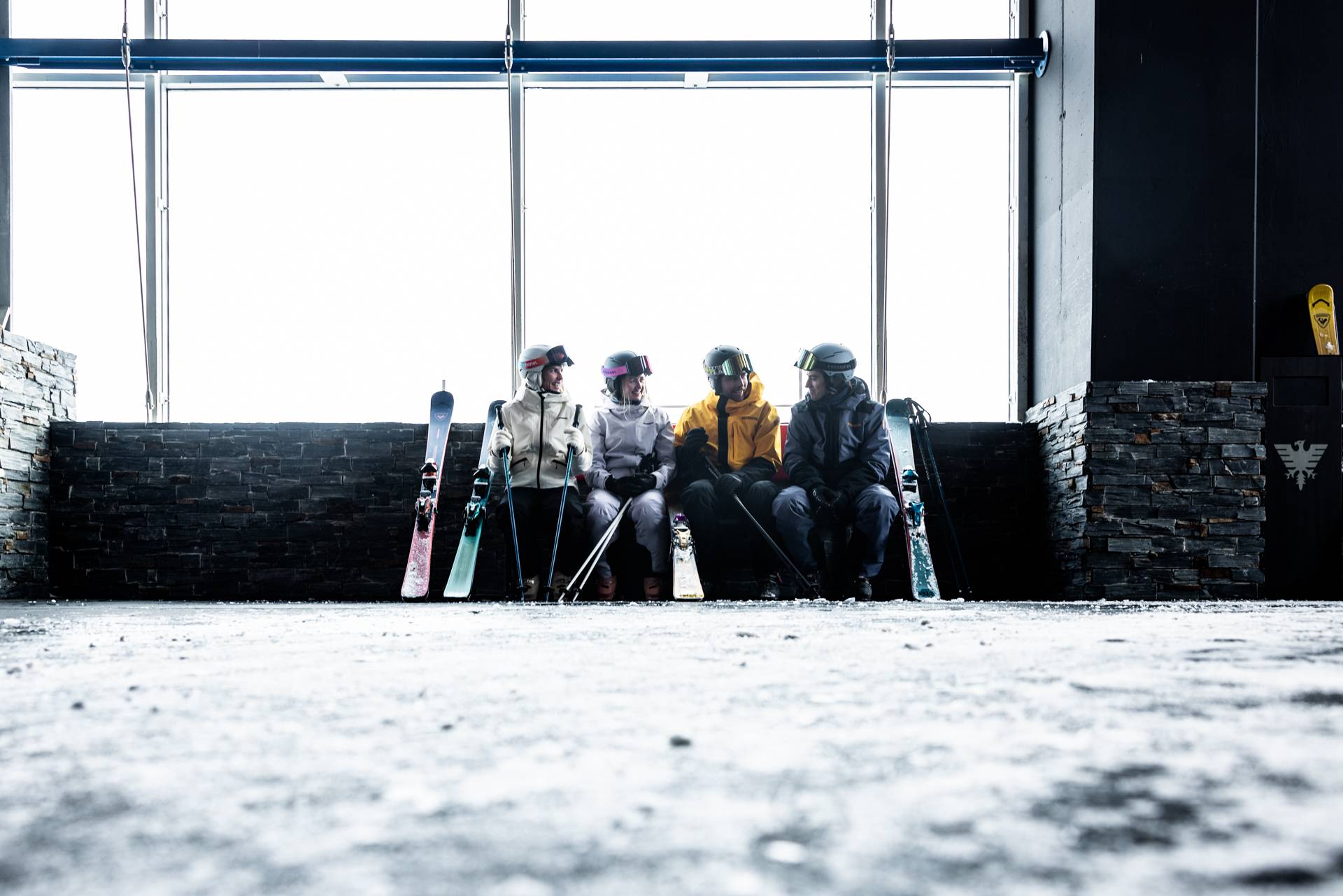 Vier personen met skiuitrusting zitten voor een gondelstation en praten met elkaar.