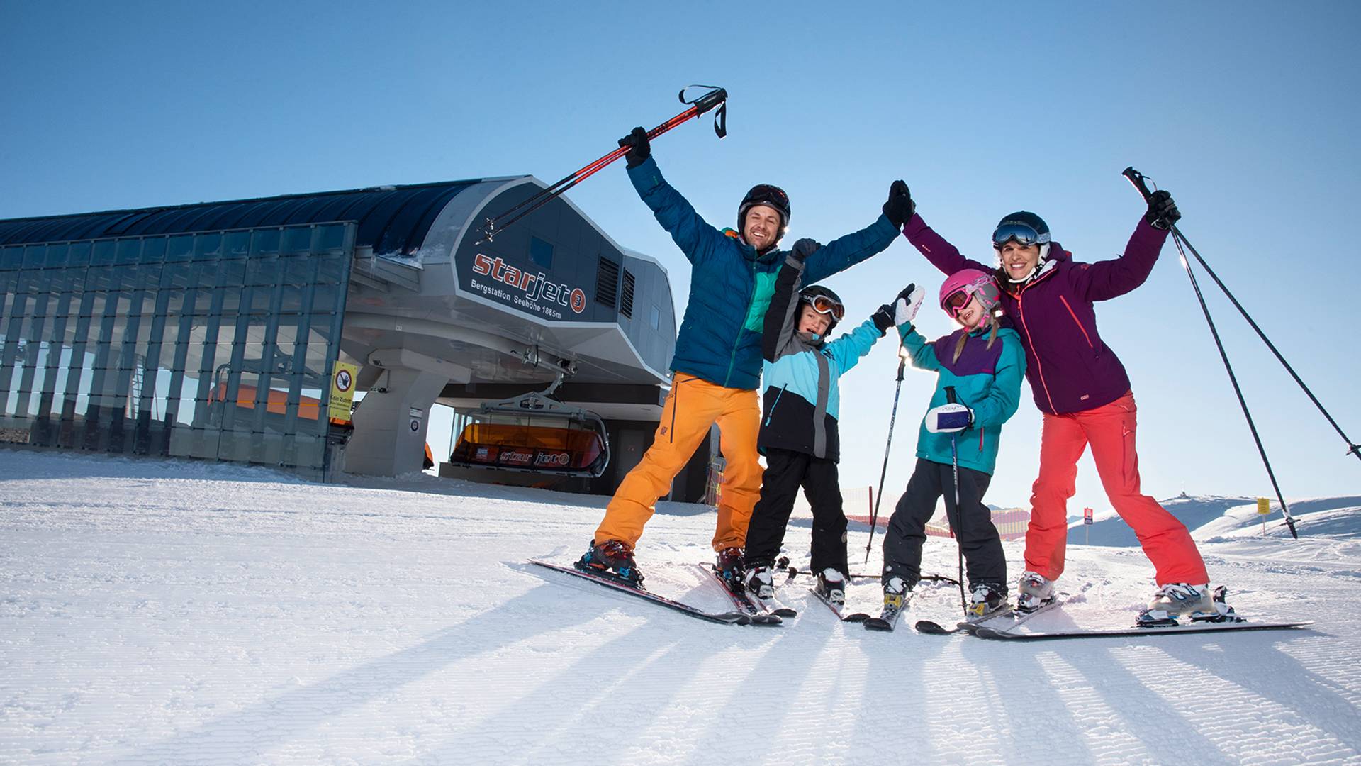 Een gezin staat voor het bergstation van een stoeltjeslift en strekt de armen omhoog.
