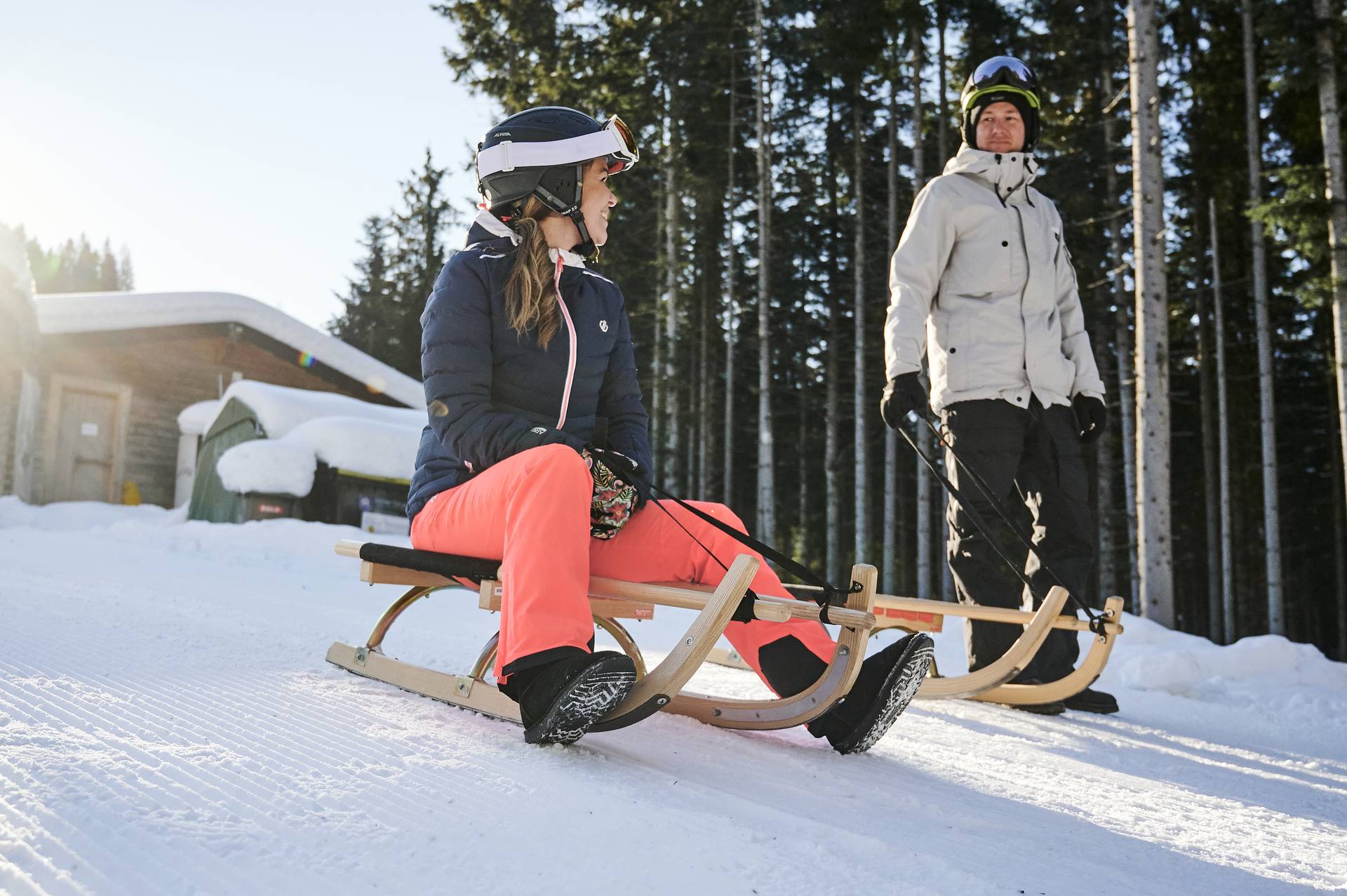 Frau sitzt auf einer Rodel, ein Mann steht neben seiner Rodel neben ihr.