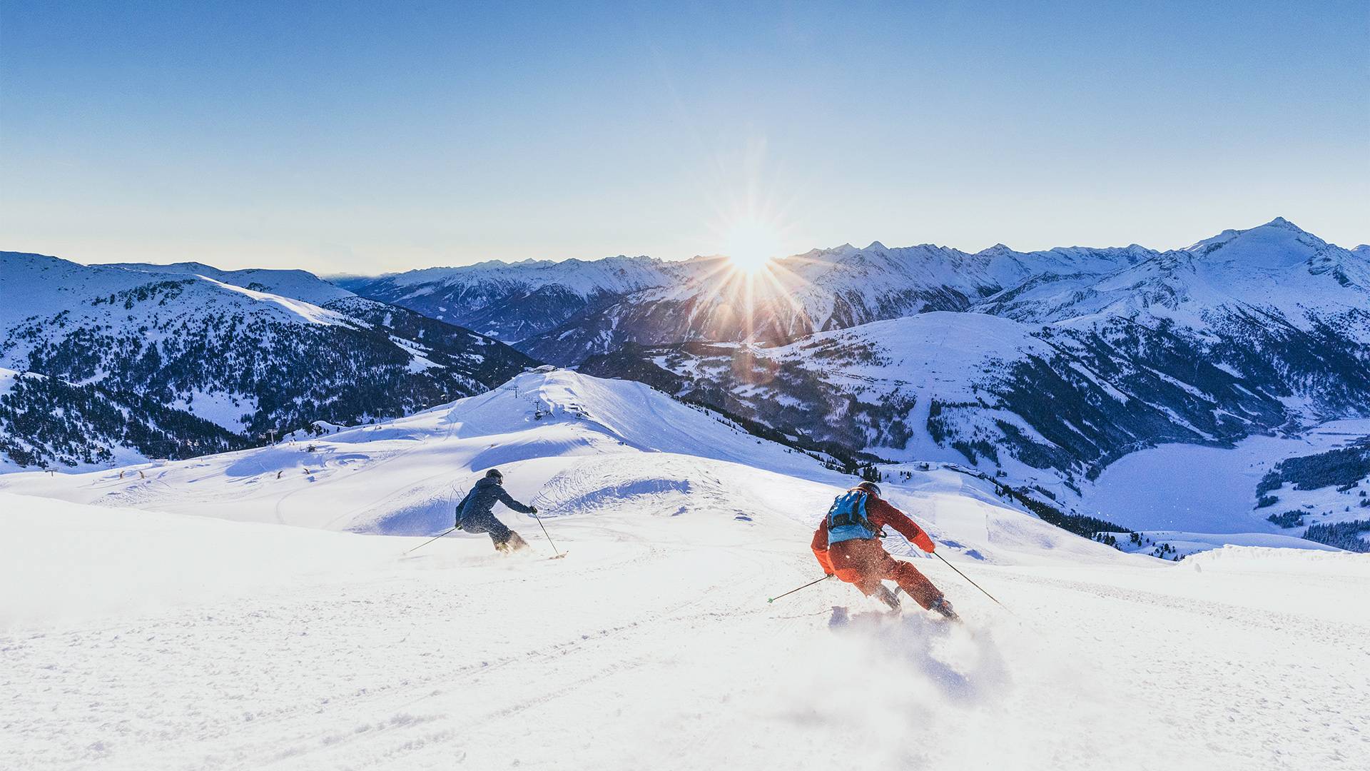 Twee personen skiën bij zonsondergang de piste af.