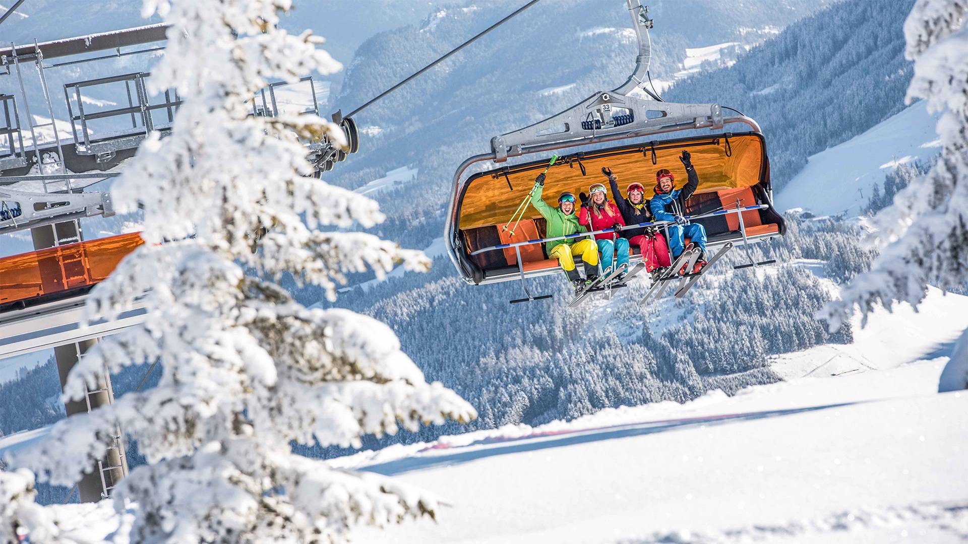 Vier Personen sitzen in einem Sessellift, im Hintergrund ist eine schneebedeckte Landschaft.