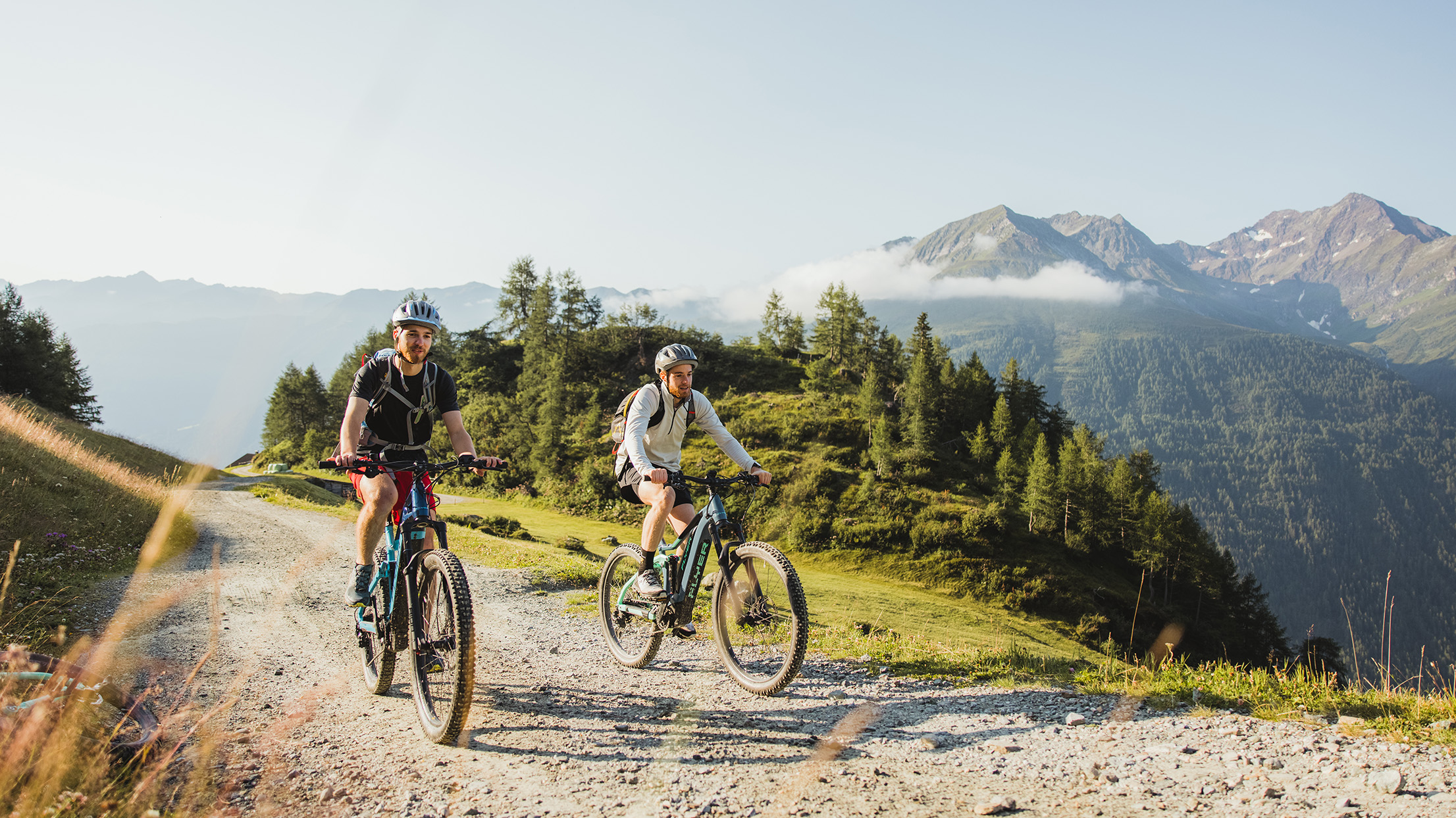 Zwei Mountainbiker fahren auf einem Berg-Radl-Weg, seitlich haben die Beiden eine herrliche Aussicht auf die schöne Ortschaft Innervillgraten.