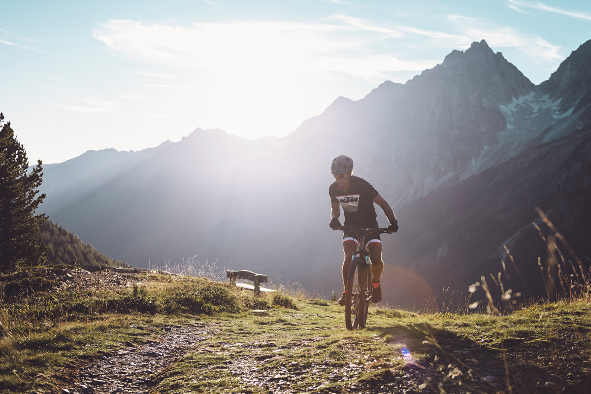 Ein Mountainbiker der gerade eine Anhöhe überwunden hat. Im Hintergrund eine abendliche Berglandschaft.