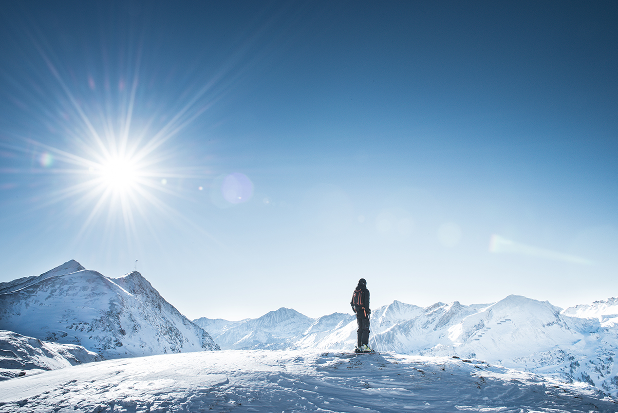 Eine Person steht auf dem Gipfel eines Berges, im Hintergrund sieht man eine Bergkette und die Sonne scheint.
